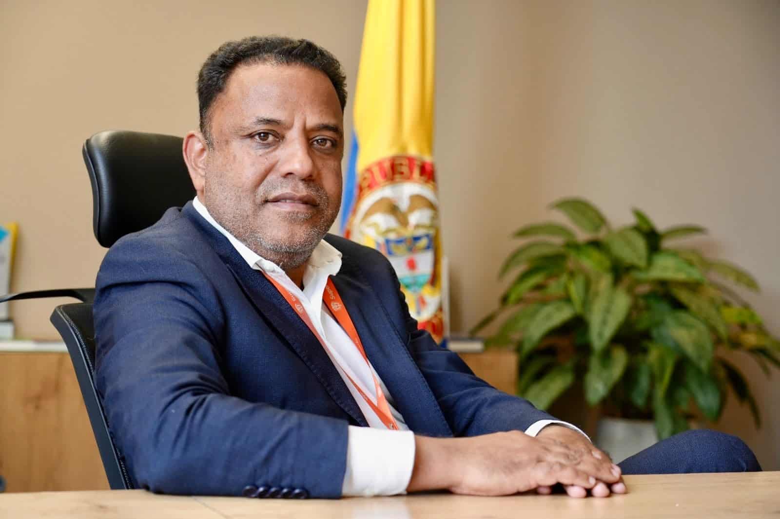 Funcionario Alfredo Enrique Piñeres Olave de la SuperTransporte sentado en oficina, con traje azul, frente a escritorio, con bandera de Colombia al fondo.