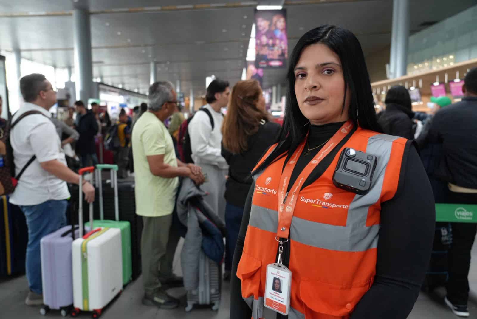 Funcionaria con chaleco de alta visibilidad en un aeropuerto, con pasajeros y equipaje en el fondo.