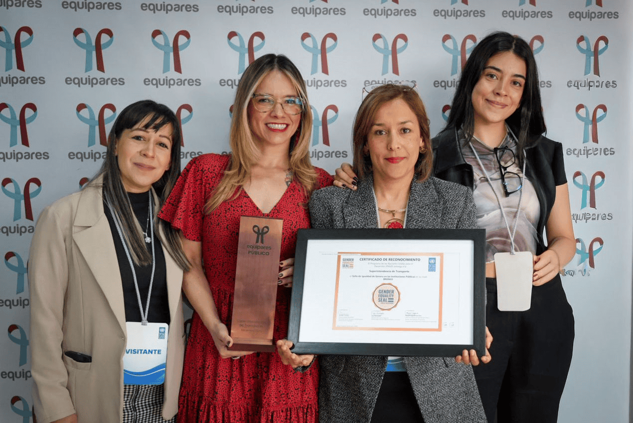 Cuatro mujeres sonríen y posan juntas en un evento institucional frente a un fondo repetido con el logo “Equipares”. En el centro, una mujer sostiene un trofeo vertical y otra un certificado enmarcado que reconoce logros en equidad de género. Todas llevan credenciales colgadas al cuello, lo que sugiere su participación en el evento.