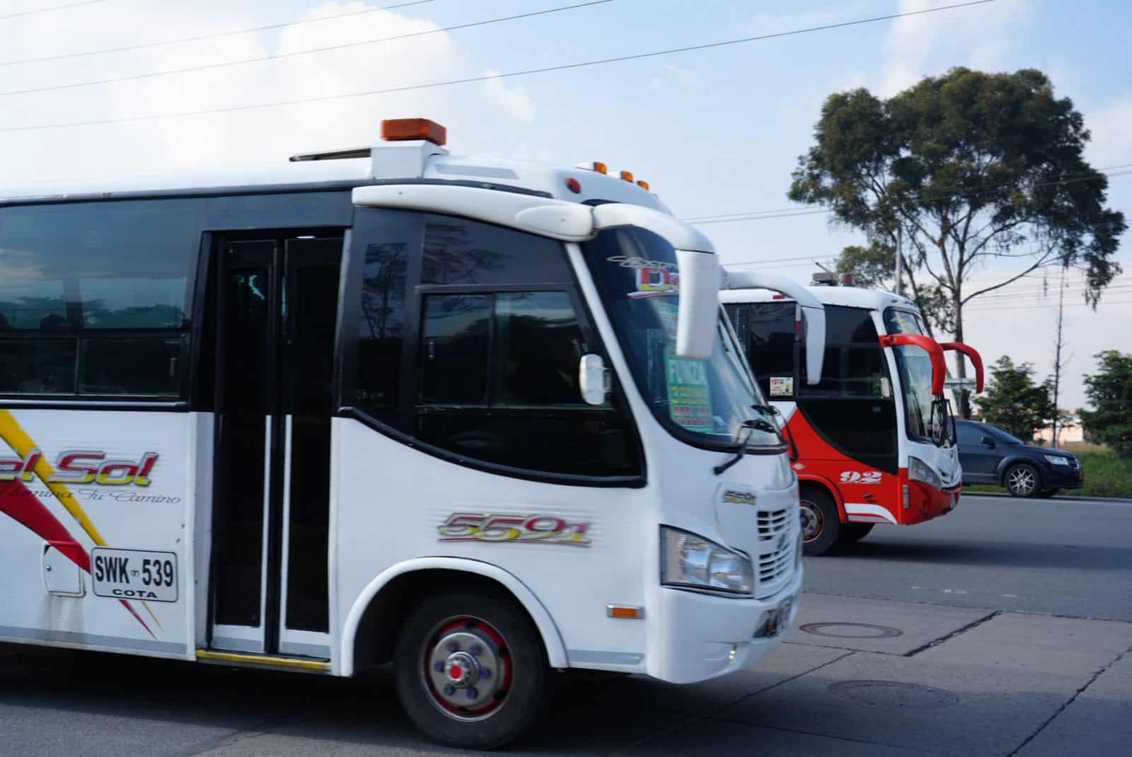 Dos buses de transporte intermunicipal circulan por una vía, uno blanco en primer plano y otro rojo detrás.