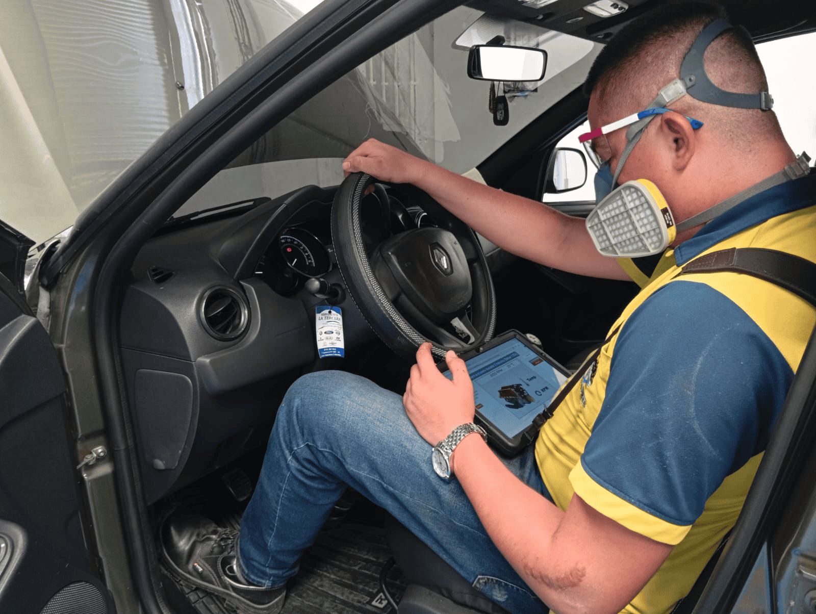 Técnico realiza inspección vehicular dentro de un automóvil usando una tableta y equipo de protección.