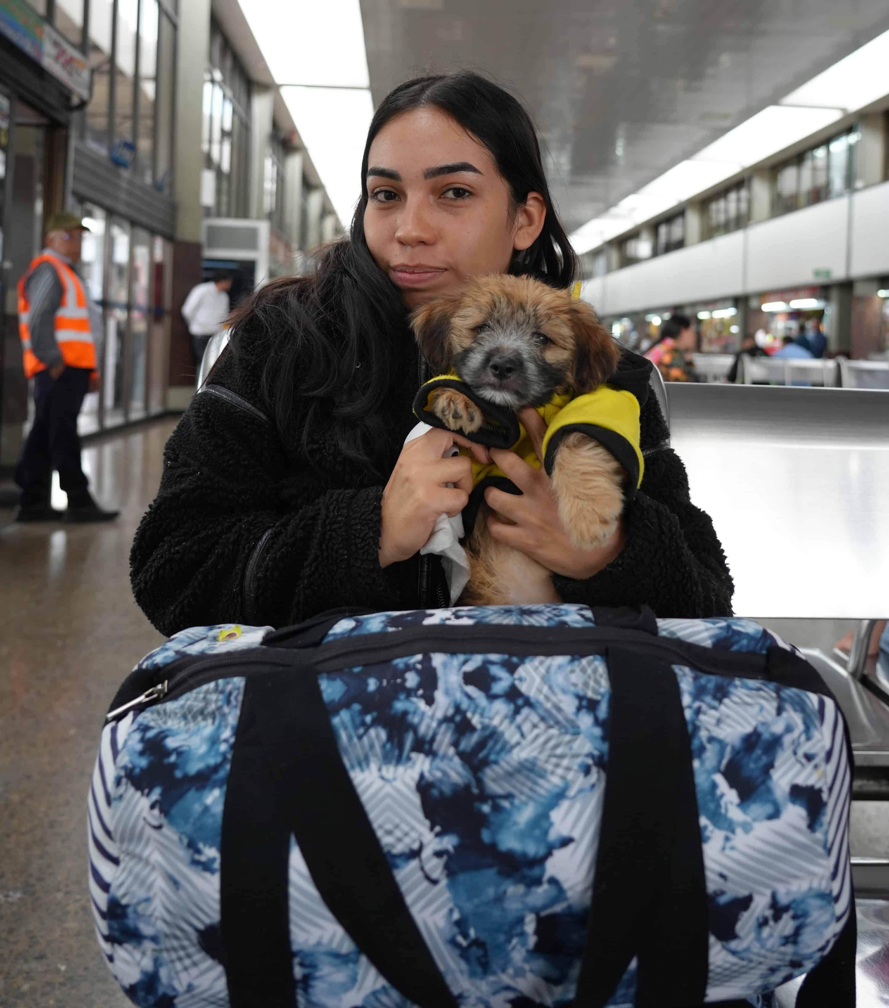 Mujer en terminal de transporte sostiene un cachorro mientras viaja con equipaje.