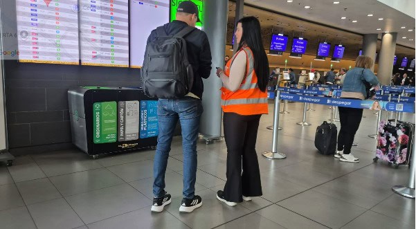 Agente de servicio al cliente de Despegar con chaleco naranja atiende a un pasajero en el aeropuerto, frente a los paneles de vuelos.