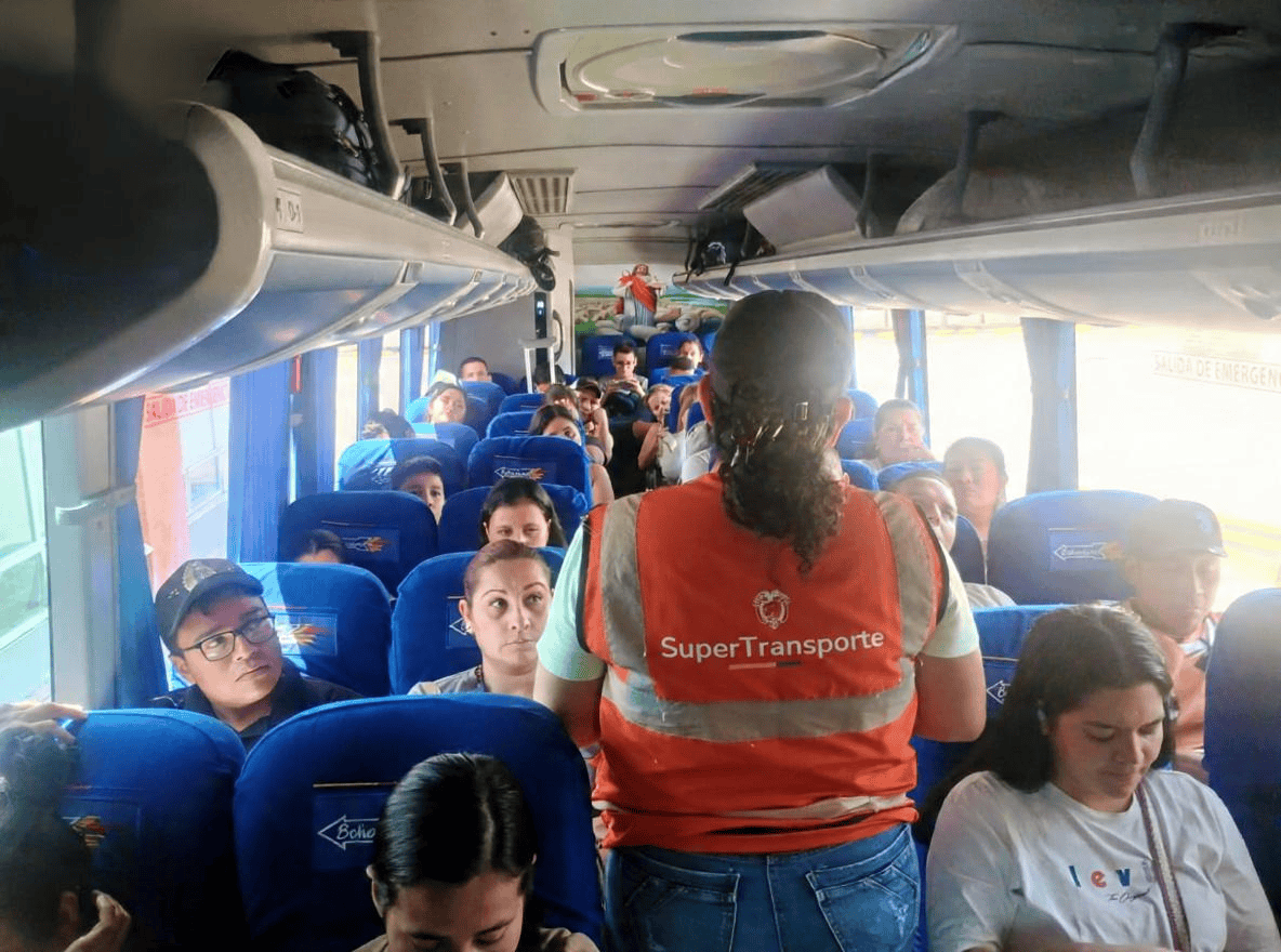 Interior de un bus lleno de pasajeros; una persona con chaleco naranja de “SuperTransporte” habla en el pasillo.
