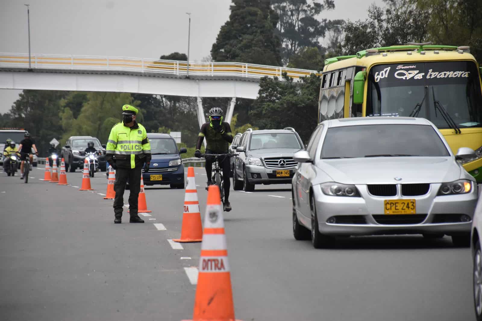 Agente de tránsito regula la circulación de vehículos y motociclistas en una vía urbana, con conos de señalización y transporte público.