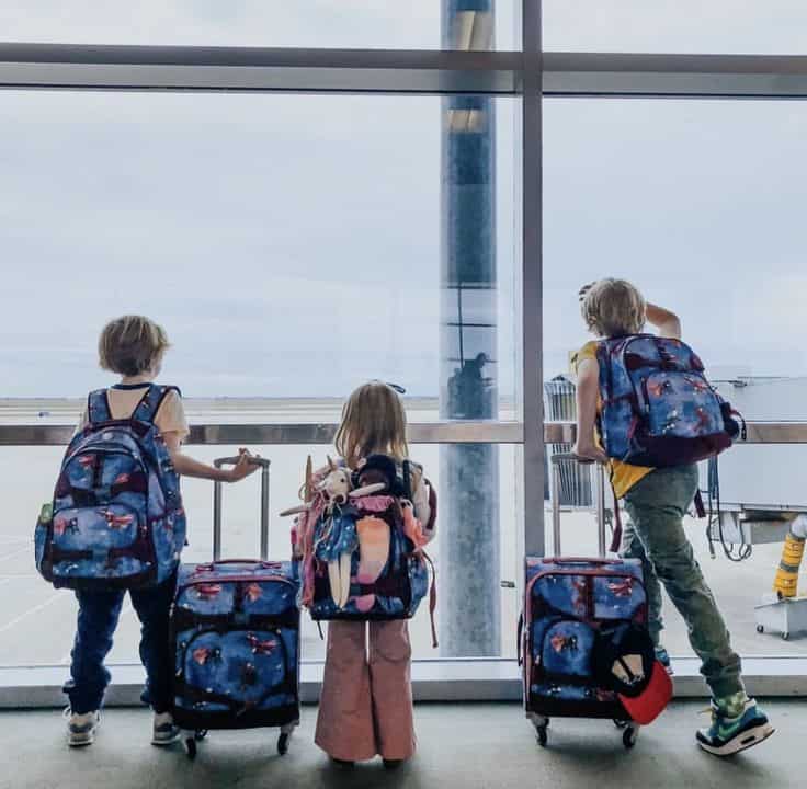 Niños con mochilas y maletas de viaje observan aviones desde la ventana de una terminal aeroportuaria.